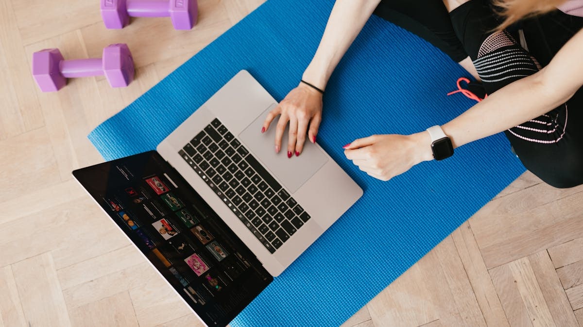 Top view of crop female in activewear sitting on yoga mat on floor and using laptop